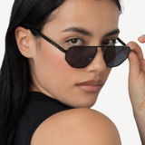 A person with long dark hair wears a sleeveless black top and tilts their head, looking over the Victoire aviator sunglasses with UV400 protection while holding the frame with one hand against a plain background.