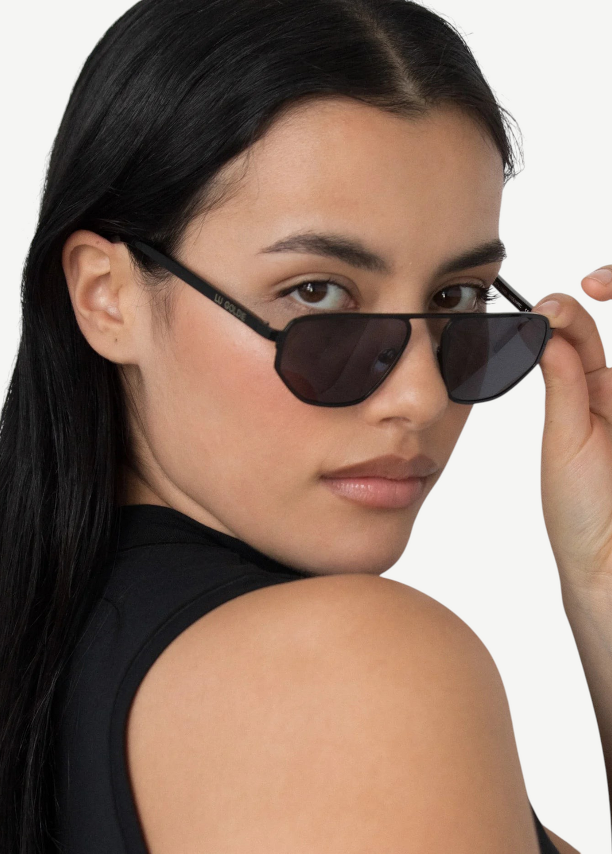 A person with long dark hair wears a sleeveless black top and tilts their head, looking over the Victoire aviator sunglasses with UV400 protection while holding the frame with one hand against a plain background.