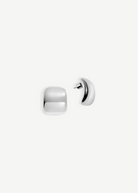 The Pebble Earrings are a pair of shiny, silver, square-shaped stud earrings with rounded edges, shown on a white background. One earring is displayed from the front, while the other reveals the side profile and pierced back. Finished with a lustrous rhodium plate, each Pebble Earring measures approximately 1 inch long.
