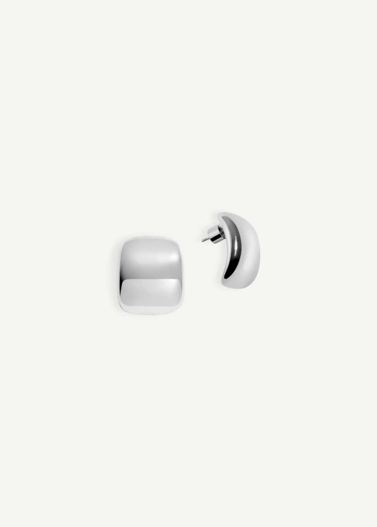 The Pebble Earrings are a pair of shiny, silver, square-shaped stud earrings with rounded edges, shown on a white background. One earring is displayed from the front, while the other reveals the side profile and pierced back. Finished with a lustrous rhodium plate, each Pebble Earring measures approximately 1 inch long.