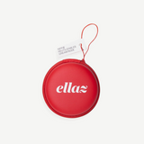 A round, red zippered case with “ellaz” printed in white letters houses Reusable Nippz nipple covers. A small attached tag reads “NIPPS, REUTILIZABLES, CREMA NUDE.” The case features a red wrist strap and is displayed against a plain white background.