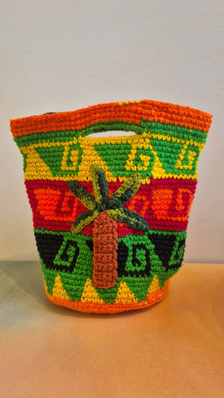 Introducing the Palmeras Bag Sac: a handmade crocheted wool bucket bag adorned with vivid geometric patterns in orange, green, yellow, and red. The front showcases a raised palm tree design, while the interior features a playful leopard lining. Set against a neutral background, this one-of-a-kind accessory energizes any setting with its vibrant style.