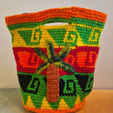 Introducing the Palmeras Bag Sac: a handmade crocheted wool bucket bag adorned with vivid geometric patterns in orange, green, yellow, and red. The front showcases a raised palm tree design, while the interior features a playful leopard lining. Set against a neutral background, this one-of-a-kind accessory energizes any setting with its vibrant style.