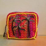 Introducing the Pochette Feliz Palmiers Fond Bag—a small, handmade crochet pouch crafted with vibrant red, yellow, and orange yarn. This eye-catching accessory features a square front panel adorned with abstract black embroidered designs and subtle leopard print-inspired accents for a unique flair. Displayed on an orange surface against a beige wall, this colorful pouch stands out as a lively addition to your collection.