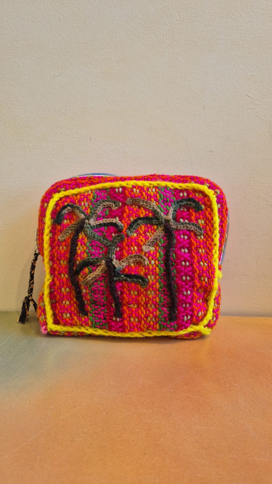 Introducing the Pochette Feliz Palmiers Fond Bag—a small, handmade crochet pouch crafted with vibrant red, yellow, and orange yarn. This eye-catching accessory features a square front panel adorned with abstract black embroidered designs and subtle leopard print-inspired accents for a unique flair. Displayed on an orange surface against a beige wall, this colorful pouch stands out as a lively addition to your collection.