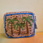 The Pochette Feliz Palmiers Fond Bag, a small, rectangular handmade pouch featuring green palm trees on a beige background with blue borders, is displayed against a cream-colored wall on an orange surface.