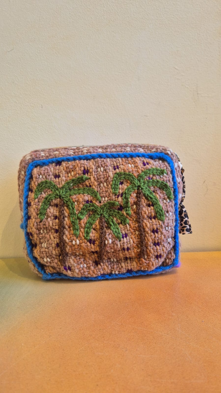 The Pochette Feliz Palmiers Fond Bag, a small, rectangular handmade pouch featuring green palm trees on a beige background with blue borders, is displayed against a cream-colored wall on an orange surface.