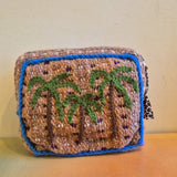 The Pochette Feliz Palmiers Fond Bag, a small, rectangular handmade pouch featuring green palm trees on a beige background with blue borders, is displayed against a cream-colored wall on an orange surface.