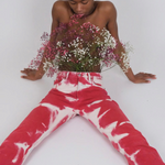 A person sits on the floor wearing vintage-style red and white Tie Dye Jeans, holding a bouquet of small pink and white flowers against their chest.