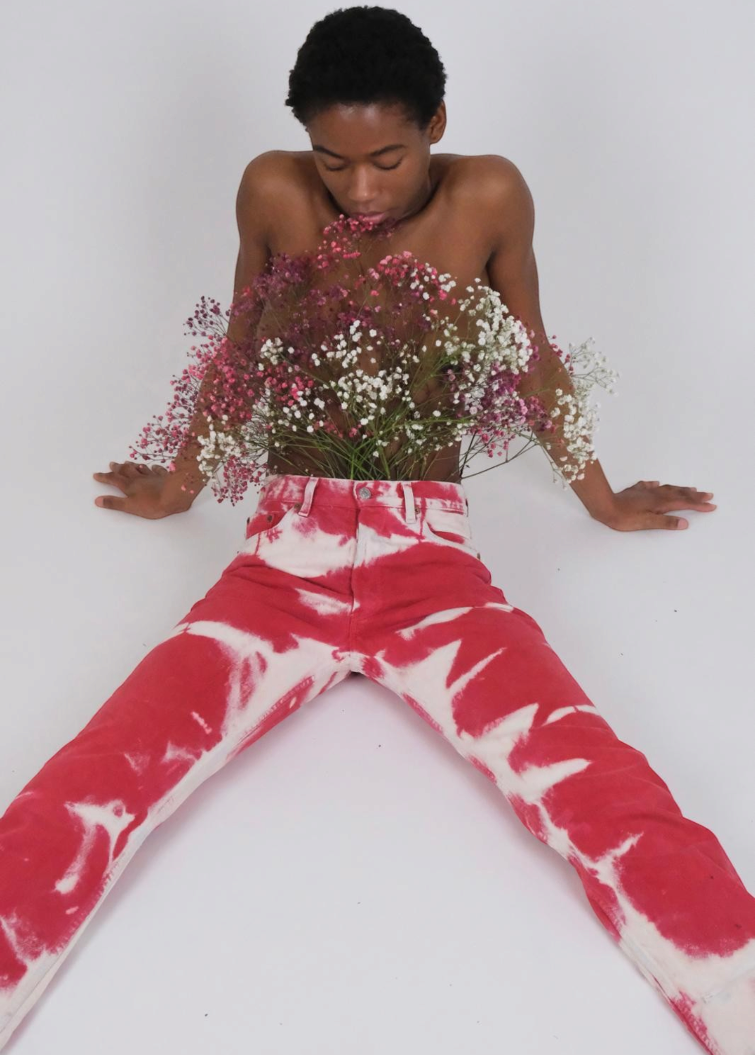 A person sits on the floor wearing vintage-style red and white Tie Dye Jeans, holding a bouquet of small pink and white flowers against their chest.