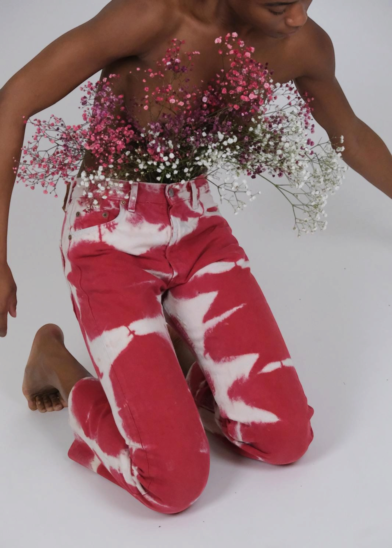 A person kneeling, wearing reworked vintage Tie Dye Jeans in red and white with a bouquet of baby’s breath tucked into the waistband.