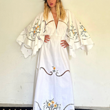 KABE SHOP INC white maxi dress with wide sleeves, floral embroidery, and cut-out details worn by a model in a hat.