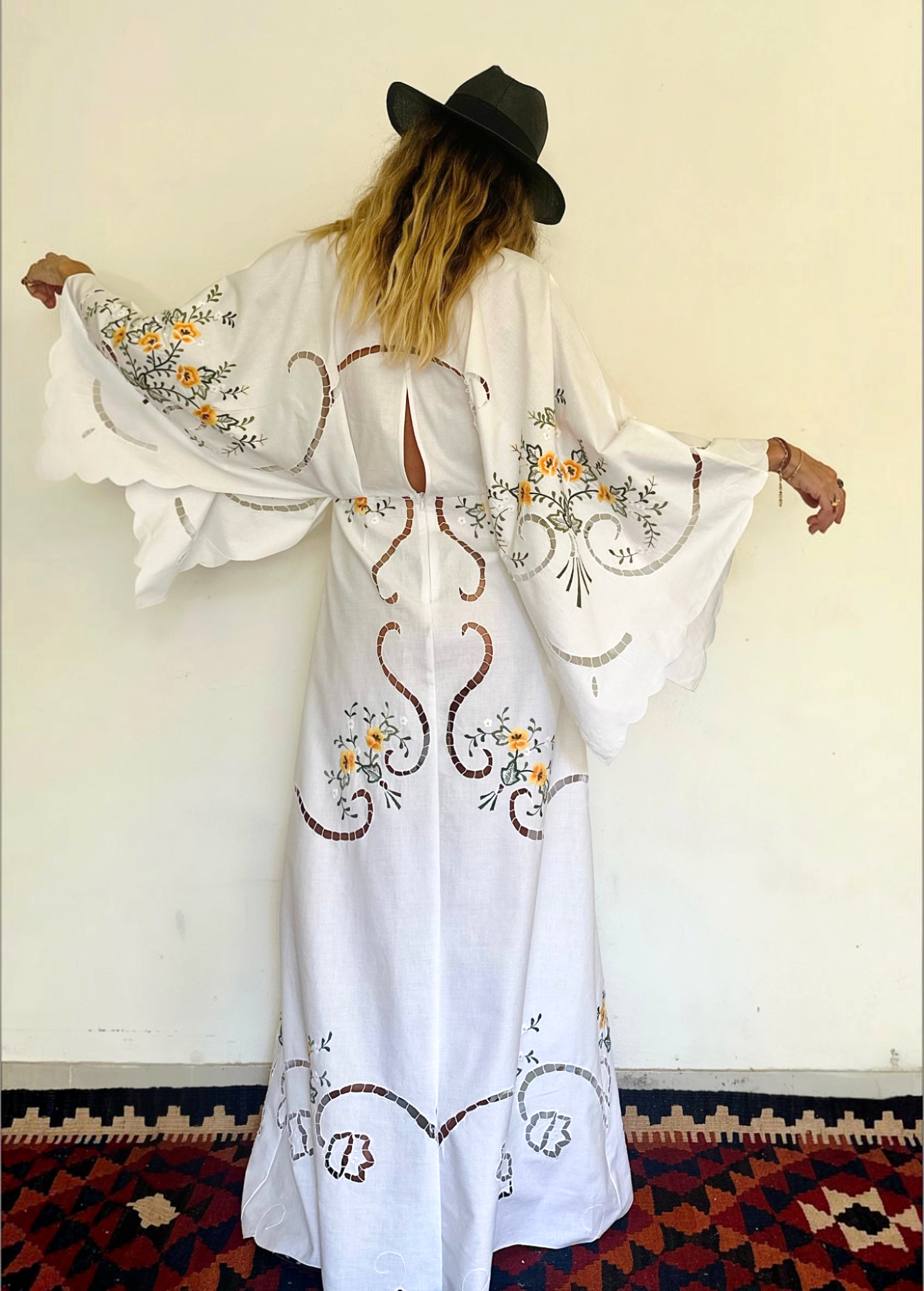 KABE SHOP INC white maxi dress with wide sleeves, floral embroidery, and cut-out details worn with a black hat.