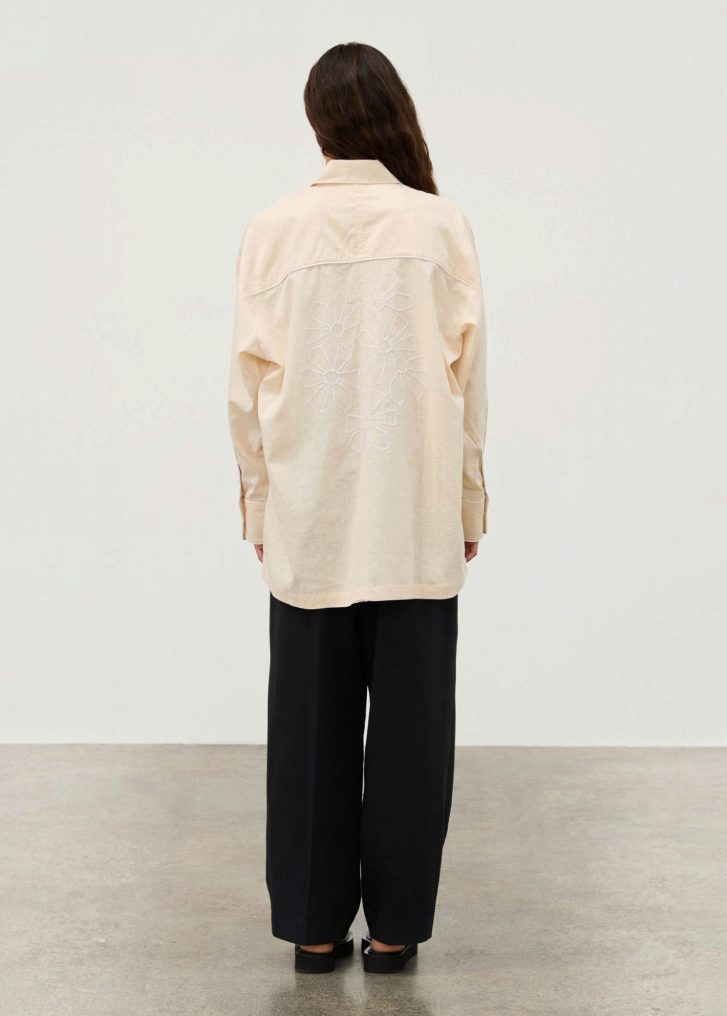 A person seen from behind wears the Shea Embroidered Hemp Blend Shirt, a long-sleeved, light-colored top with white geometric floral embroidery on the back, paired with loose black trousers.