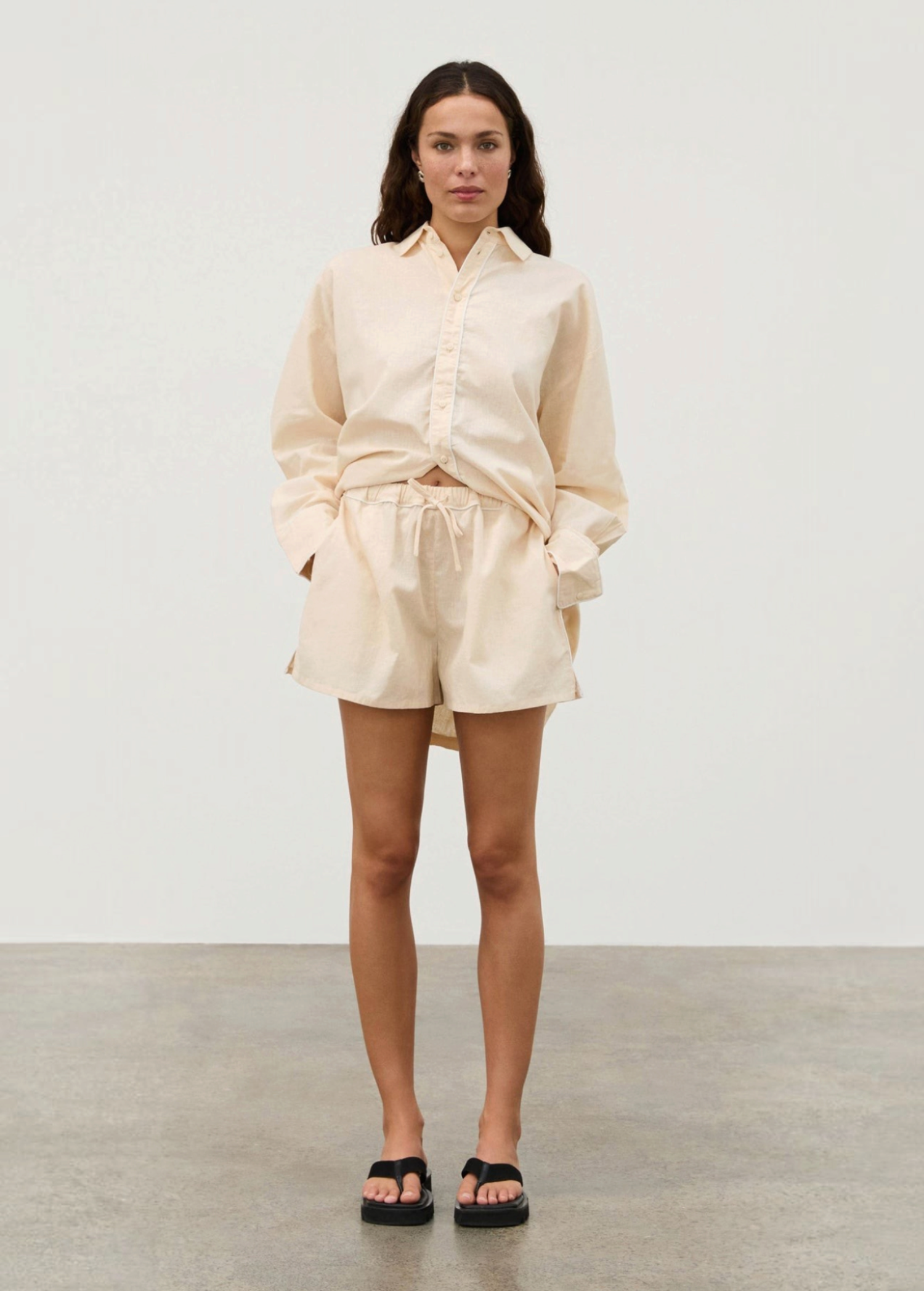 A woman wearing a beige long-sleeved Shea Embroidered Hemp Blend Shirt with geometric floral detailing, matching drawstring shorts, and black sandals.