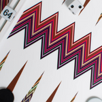 A close-up of the ZigZag Backgammon Board Game, featuring a vibrant acrylic zigzag design with black and white checkers, a doubling cube set to 64, and a white die displaying two dots. The bold zigzag patterns make this board an eye-catching addition to any coffee table.
