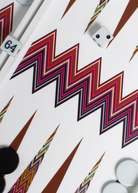 A close-up of the ZigZag Backgammon Board Game, featuring a vibrant acrylic zigzag design with black and white checkers, a doubling cube set to 64, and a white die displaying two dots. The bold zigzag patterns make this board an eye-catching addition to any coffee table.