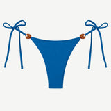 Praia Bottom in blue, crafted from eco-friendly Peach Perfect fabric, features adjustable side tie strings with brown bead accents on each hip. Displayed on a white background, this swimwear combines comfort and style for a flawless fit.
