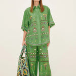 A person wears a Sun Dancer Shirt and matching wide-leg trousers in a classic silhouette, both featuring colorful embroidered folk-art motifs, and carries a large patterned tote bag.