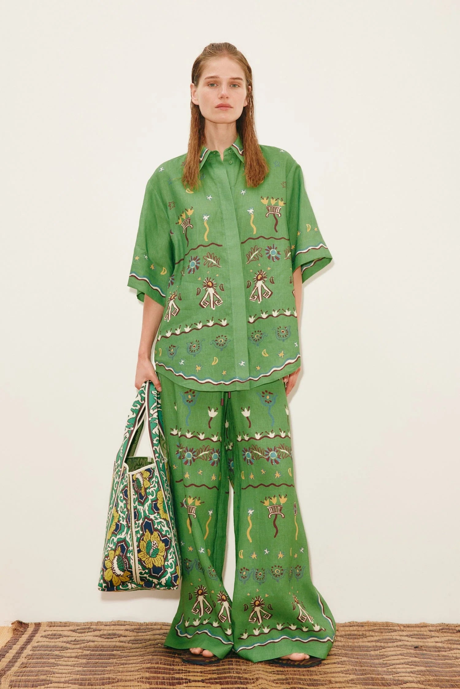 A person wears a Sun Dancer Shirt and matching wide-leg trousers in a classic silhouette, both featuring colorful embroidered folk-art motifs, and carries a large patterned tote bag.