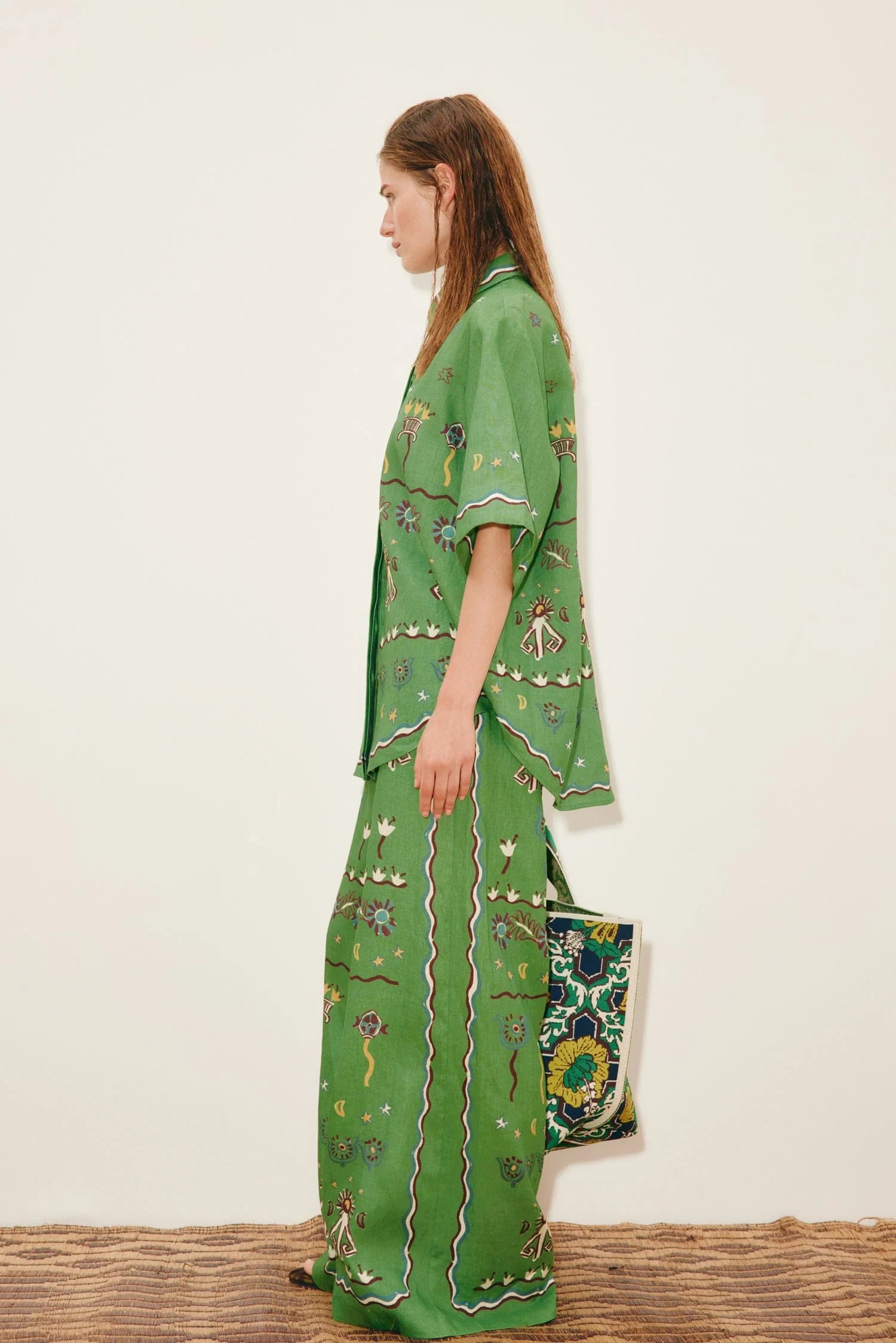 A person wears a Sun Dancer Shirt and matching wide-leg trousers, both decorated with colorful floral and geometric patterns, while holding a matching tote bag to complete the classic silhouette.