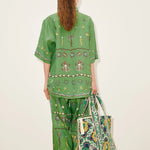 A person seen from behind wears a Sun Dancer Shirt and matching wide-leg trousers in green, both decorated with embroidered flowers, suns, and abstract shapes, while carrying a large patterned tote bag.