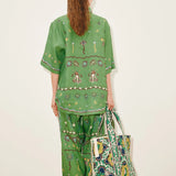 A person seen from behind wears a Sun Dancer Shirt and matching wide-leg trousers in green, both decorated with embroidered flowers, suns, and abstract shapes, while carrying a large patterned tote bag.