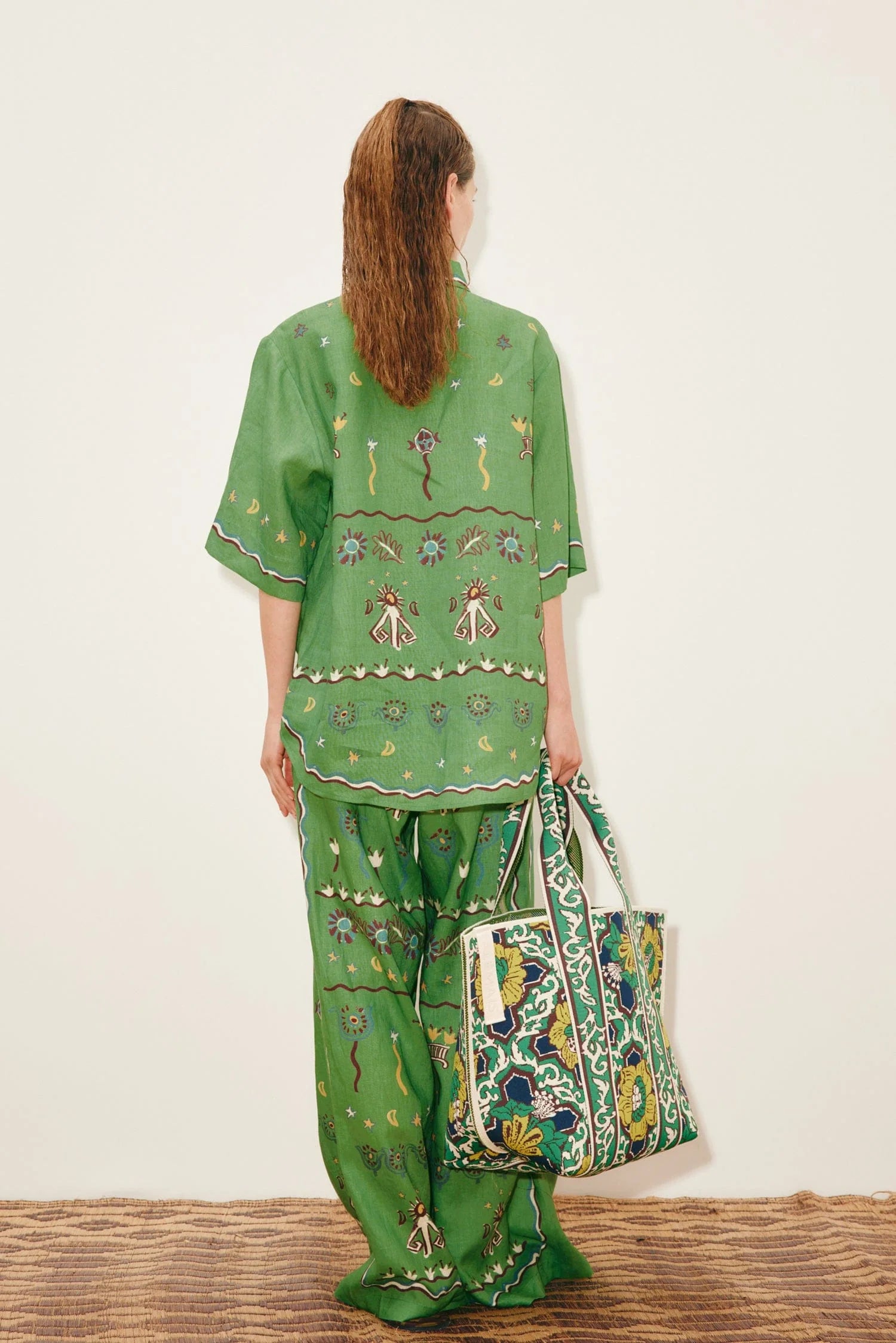 A person seen from behind wears a Sun Dancer Shirt and matching wide-leg trousers in green, both decorated with embroidered flowers, suns, and abstract shapes, while carrying a large patterned tote bag.