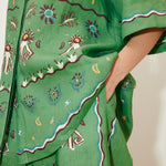 Sun Dancer Shirt: A green, short-sleeved, relaxed-fit button-up featuring colorful embroidered sun, star, and floral patterns, with a hand in the pocket.