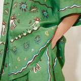 Sun Dancer Shirt: A green, short-sleeved, relaxed-fit button-up featuring colorful embroidered sun, star, and floral patterns, with a hand in the pocket.