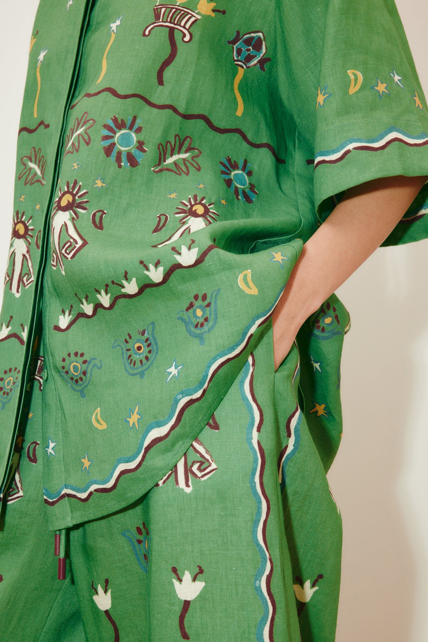 Sun Dancer Shirt: A green, short-sleeved, relaxed-fit button-up featuring colorful embroidered sun, star, and floral patterns, with a hand in the pocket.