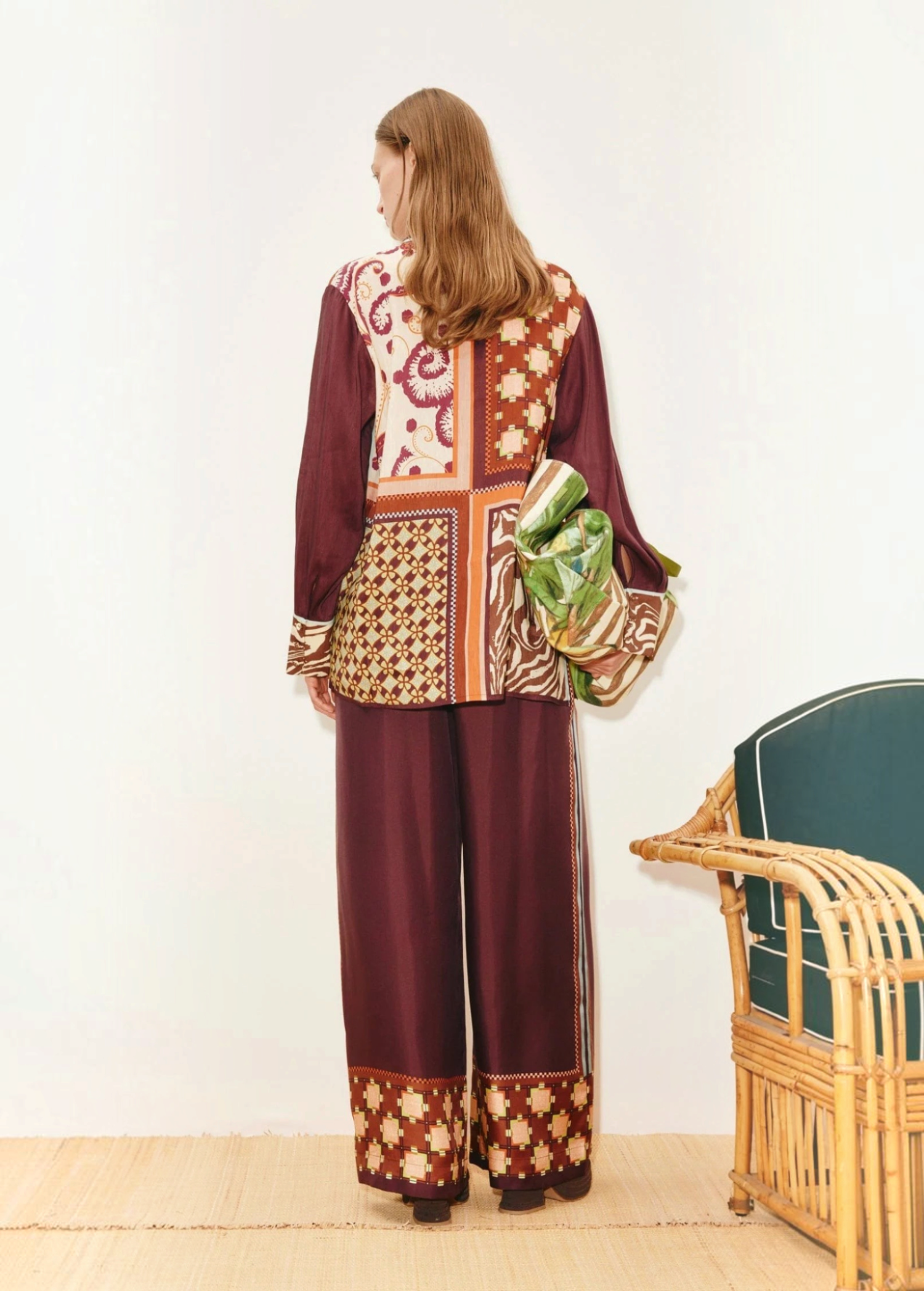 A person wears a Wilson Shirt and matching wide-leg trousers, both featuring maroon-toned panels with varied geometric and floral patterns, while holding a green patterned bag.
