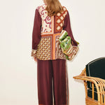 A person wears a Wilson Shirt and matching wide-leg trousers, both featuring maroon-toned panels with varied geometric and floral patterns, while holding a green patterned bag.