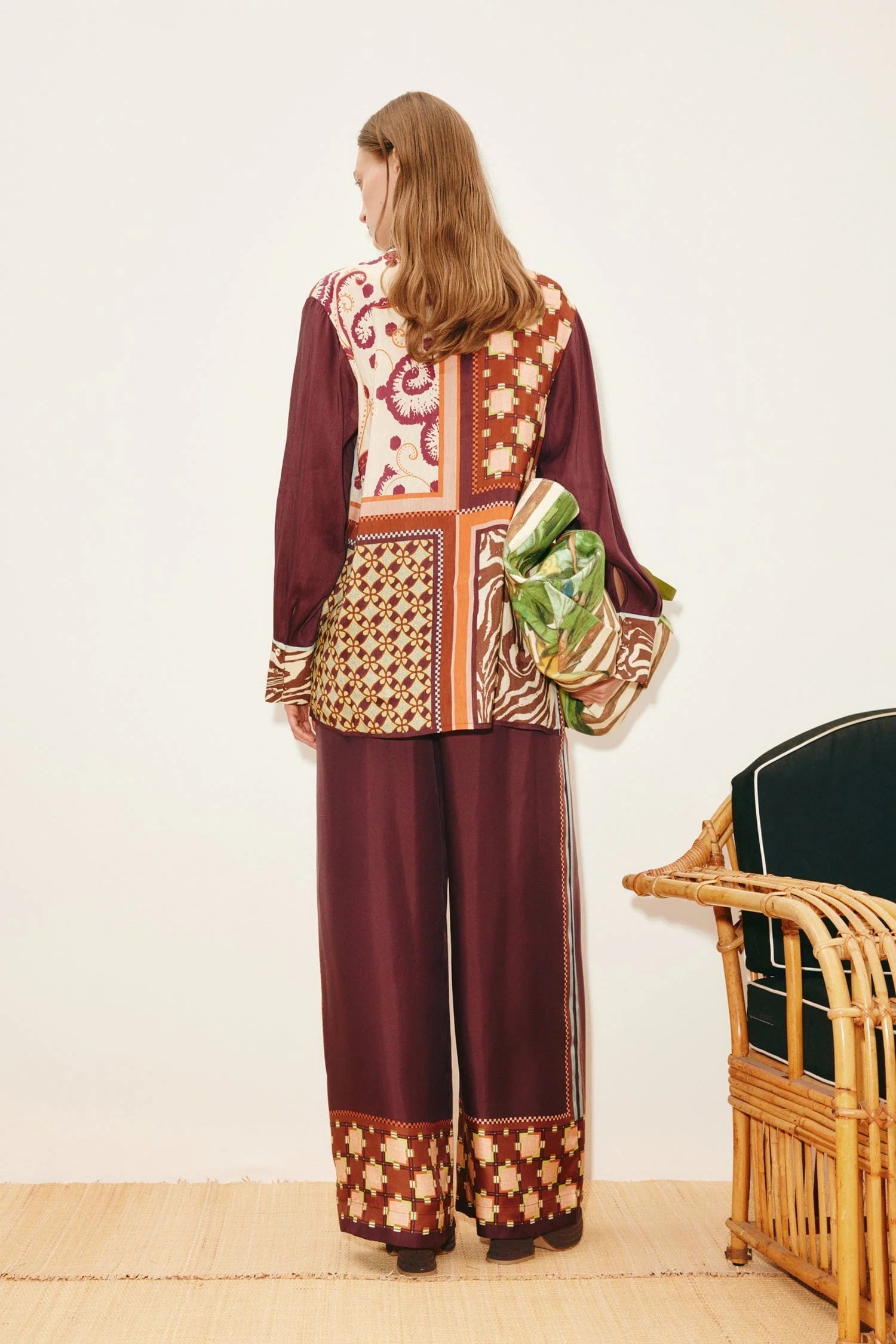 A person wears a Wilson Shirt and matching wide-leg trousers, both featuring maroon-toned panels with varied geometric and floral patterns, while holding a green patterned bag.