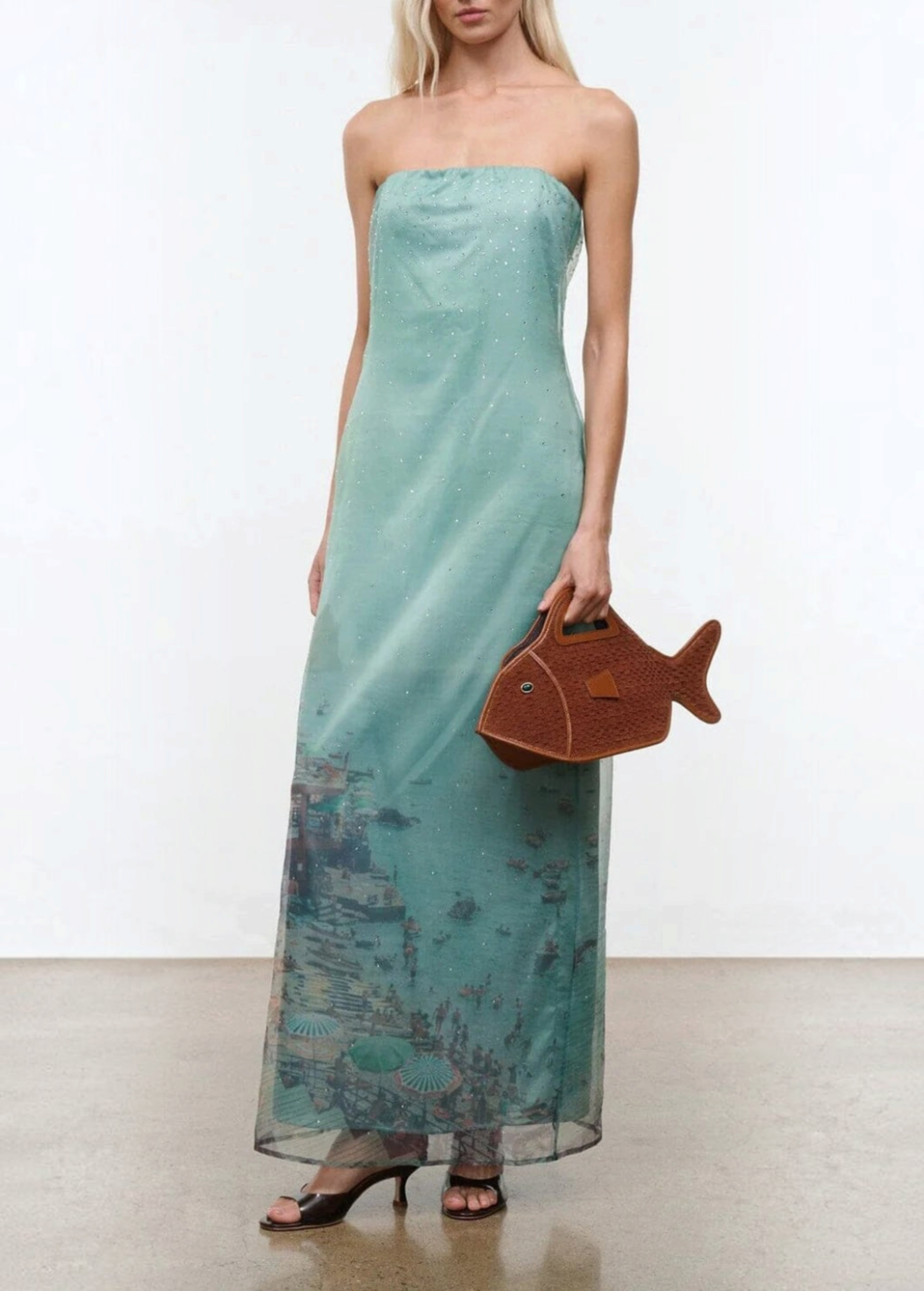 Light green strapless floor-length Amara Dress with a graphic beach scene print at the hem, styled with black heeled sandals and a brown fish-shaped handbag for special occasions.