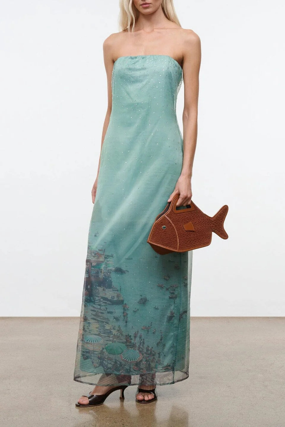 Light green strapless floor-length Amara Dress with a graphic beach scene print at the hem, styled with black heeled sandals and a brown fish-shaped handbag for special occasions.