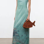 Light green strapless floor-length Amara Dress with a graphic beach scene print at the hem, styled with black heeled sandals and a brown fish-shaped handbag for special occasions.