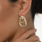 Close-up of a woman’s ear showcasing the Ami Earrings: chunky, 1.5-inch drop double hoops in a striking 14KT gold plate with a pierced back. Her hand gently touches her neck, while her dark hair is pulled away to reveal the bold gold-tone jewelry.