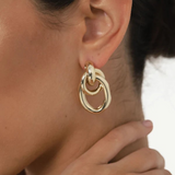 Close-up of a woman’s ear showcasing the Ami Earrings: chunky, 1.5-inch drop double hoops in a striking 14KT gold plate with a pierced back. Her hand gently touches her neck, while her dark hair is pulled away to reveal the bold gold-tone jewelry.
