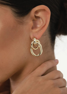 Close-up of a woman’s ear showcasing the Ami Earrings: chunky, 1.5-inch drop double hoops in a striking 14KT gold plate with a pierced back. Her hand gently touches her neck, while her dark hair is pulled away to reveal the bold gold-tone jewelry.