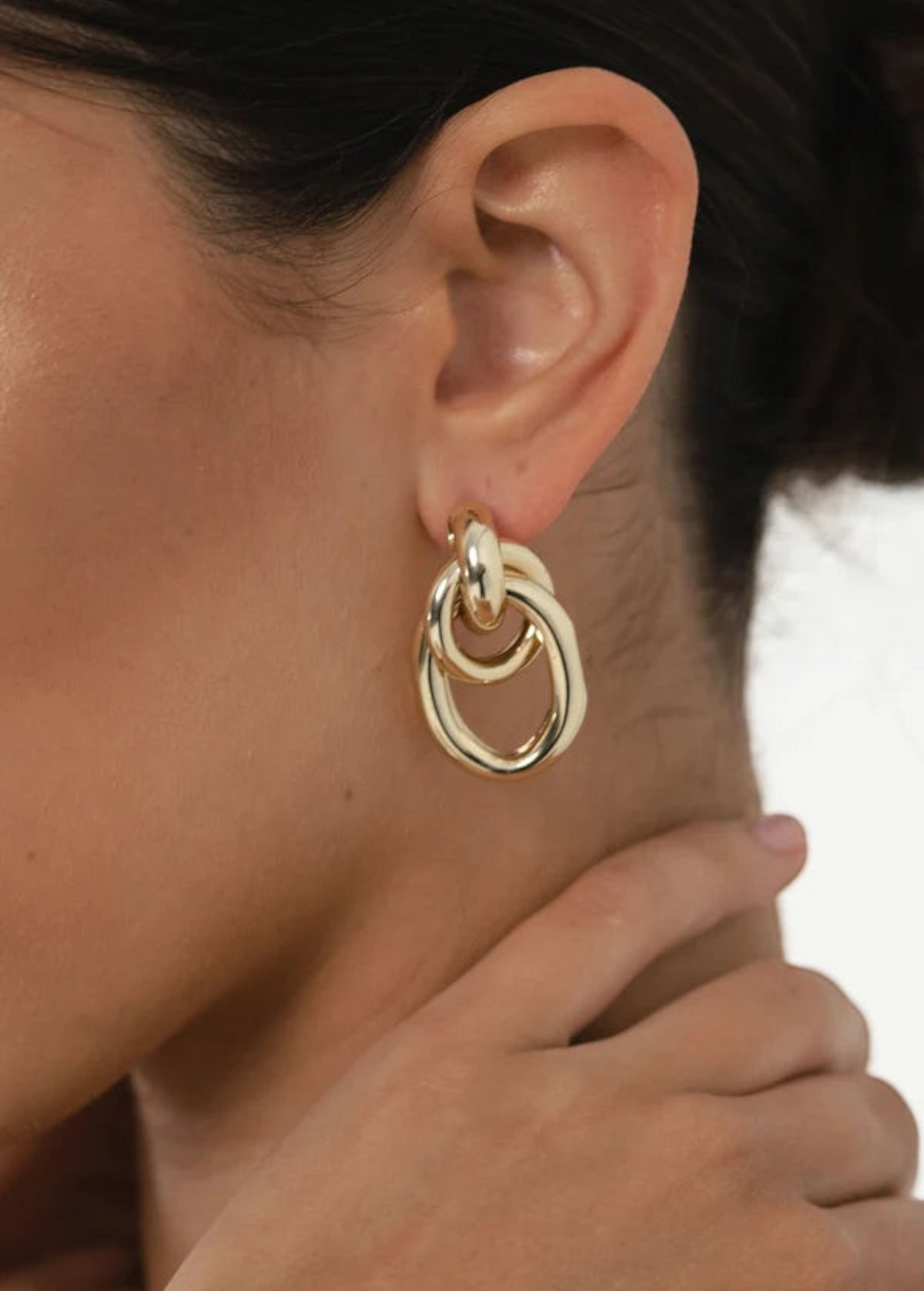 Close-up of a woman’s ear showcasing the Ami Earrings: chunky, 1.5-inch drop double hoops in a striking 14KT gold plate with a pierced back. Her hand gently touches her neck, while her dark hair is pulled away to reveal the bold gold-tone jewelry.