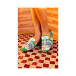 Wearing the Anzu Moccasin—colorful, beaded green footwear featuring Egyptian-inspired patterns and a babouche-style silhouette—a person stands on a red and white tiled floor set against a warm, textured backdrop.
