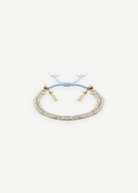 The N° 183 Braided Bracelet, crafted from luxurious amethyst silk yarn in light blue and gold threads with a radiant yellow gold finish, features elegant gold bead accents and adjustable tassel ends. It is displayed on a plain white background.