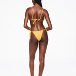 A woman with braided hair stands facing away, wearing an eco-friendly yellow Praia Bottom with adjustable side ties and black sandals, against a plain white background.