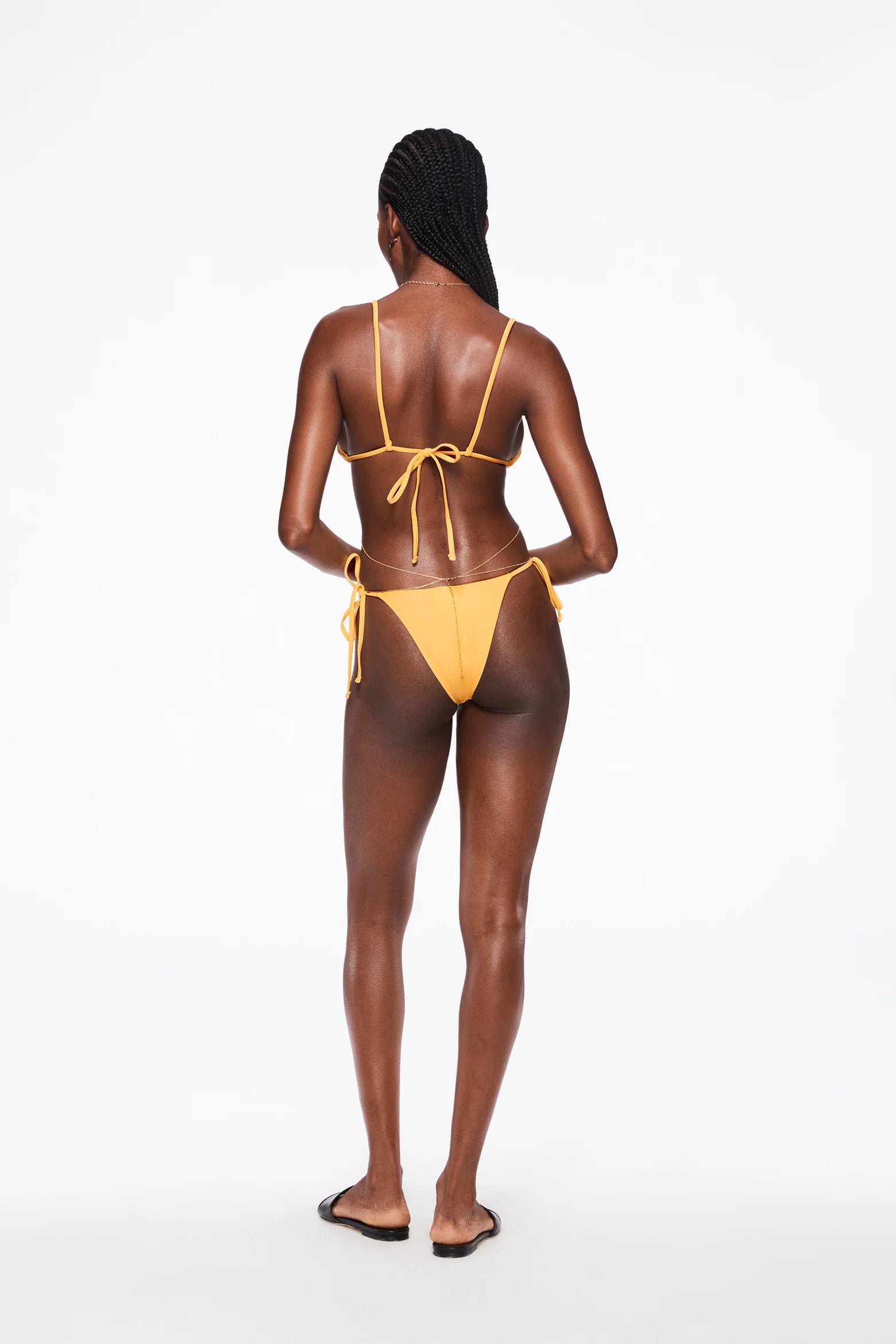 A woman with braided hair stands facing away, wearing an eco-friendly yellow Praia Bottom with adjustable side ties and black sandals, against a plain white background.