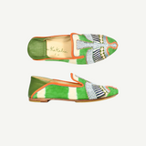 Two green and orange Anzu Moccasin shoes with intricate hand-embroidered beaded designs on the toes, displayed on a white background. The tan insoles complement geometric and wing-like patterns in white, blue, and black beadwork. These elegant slip-ons showcase exquisite moccasin craftsmanship for a truly unique finish.