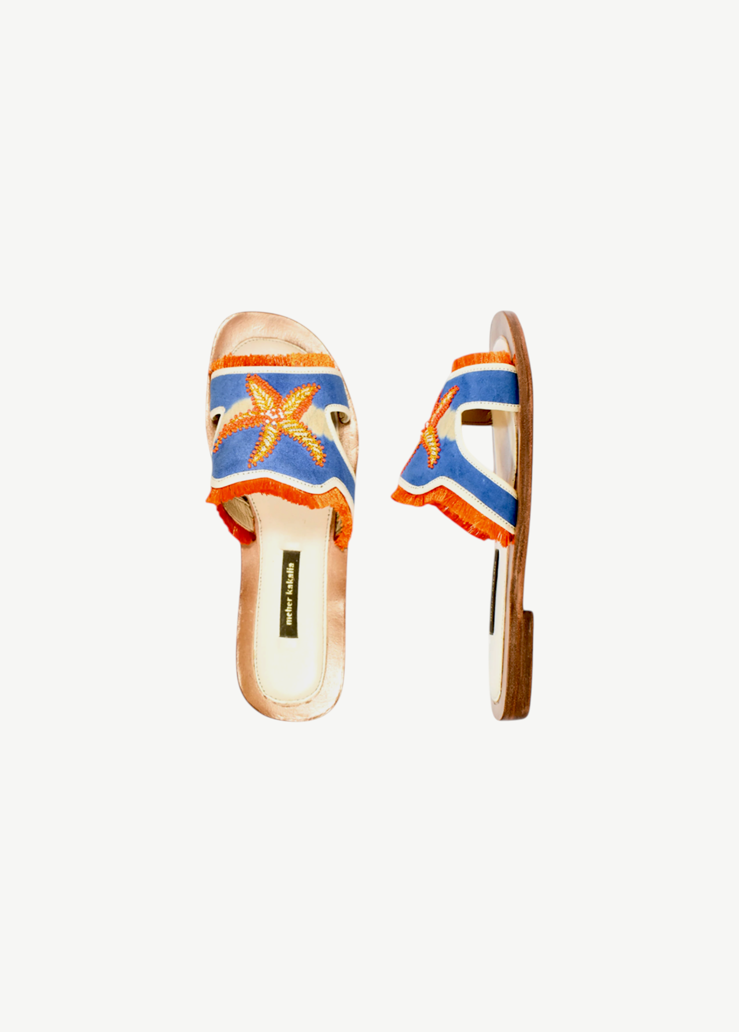 Introducing the Mazu Slide: a pair of hand-embroidered leather slip-on sandals featuring blue and orange accents, playful fringe trim, and a decorative starfish adorned with shell and bead details on the upper strap. Showcasing Silk Route’s artisan craftsmanship, these slides blend unique textures with coastal-inspired embellishments for a truly distinctive look.