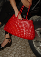 Seated in a car, an individual holds the Santa Croce Big Bag—a striking handwoven leather tote whose vibrant red hue catches the light. They wear black pants and black wedge heels, with one foot resting on the cobblestone surface beside the car tire.