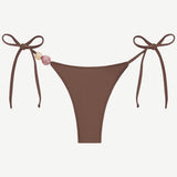 Praia Bottom in brown with adjustable tie sides, adorned with a pink and a beige bead on the left string, crafted from recycled polyamide for an eco-friendly swimwear option, shown on a white background.