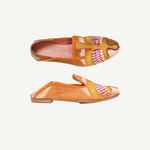 Two orange Anzu Moccasins featuring intricate pink, yellow, and gold beaded embroidery on the uppers are displayed side by side on a white background. These hand-embroidered moccasins showcase artful craftsmanship and style, making them a unique statement piece.
